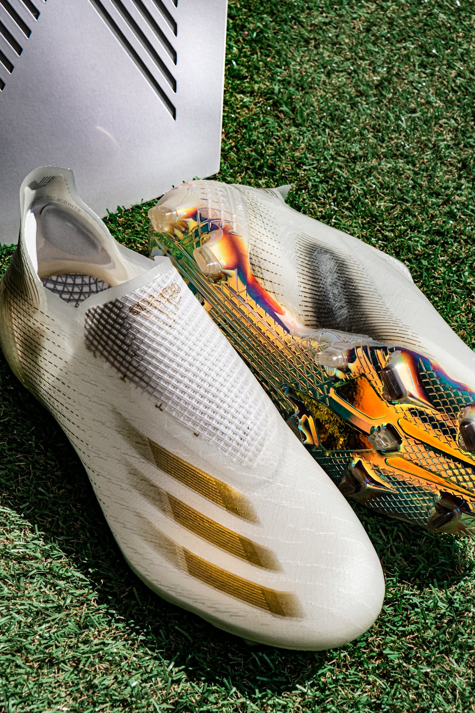 Two white soccer cleats on green turf with gold accents.