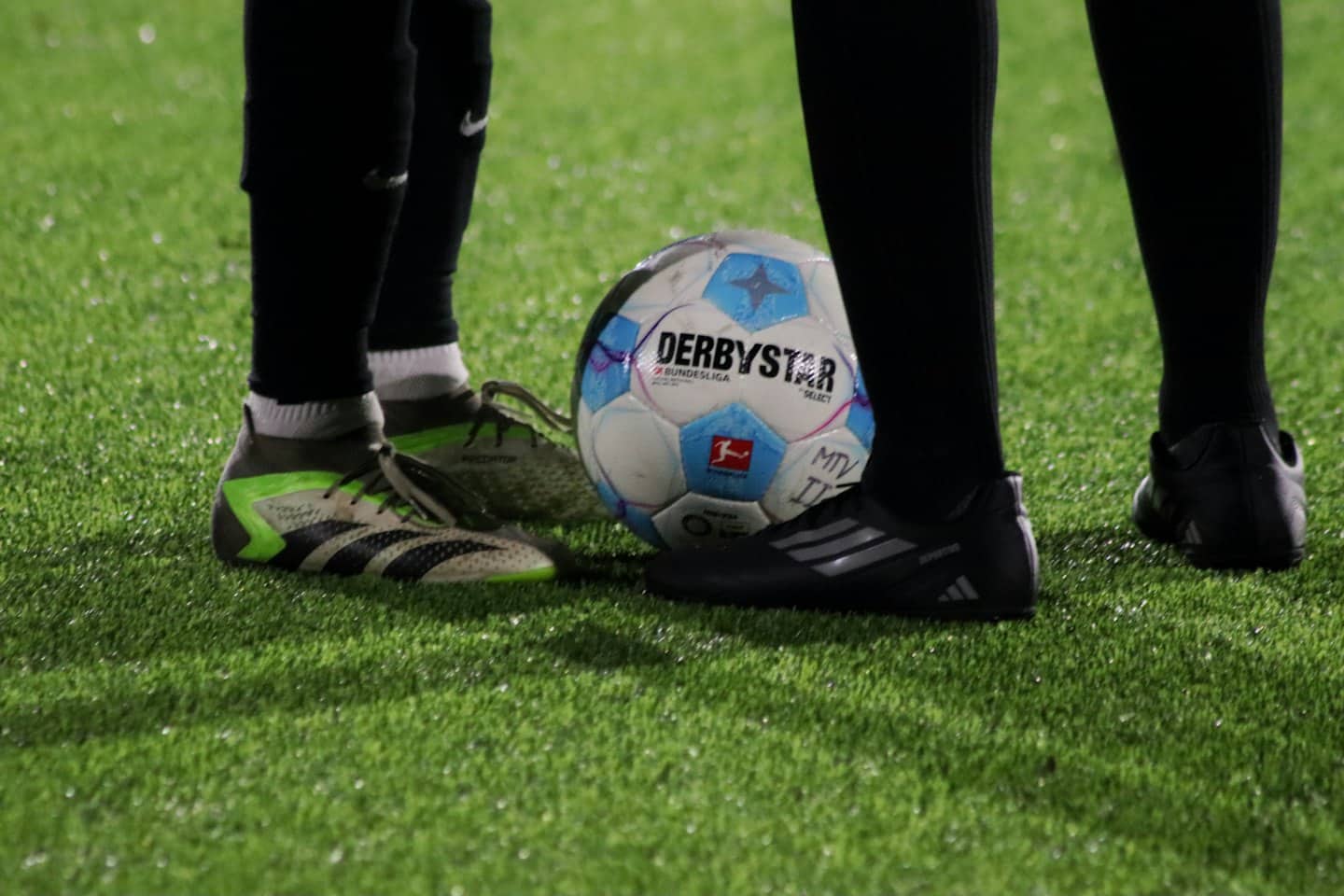 Two soccer players stand on artificial turf with a ball.